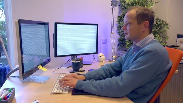 Man working in an office behind a computer witth two screens typing frantically on a keyboard with ten fingers