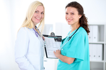 Beautiful young smiling female doctor standing at hospital with doctor on the background. Medical concept.