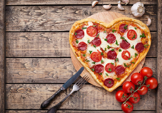 Heart shaped pizza with pepperoni, tomatoes and mozzarella on vintage wooden table background. Valentines day love concept. 