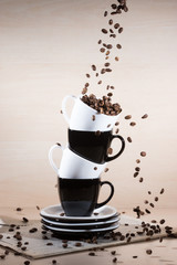 View of black and white cups on the stack of the plates with falling down brown roasted coffee beans on newspaper on light wooden textured background.