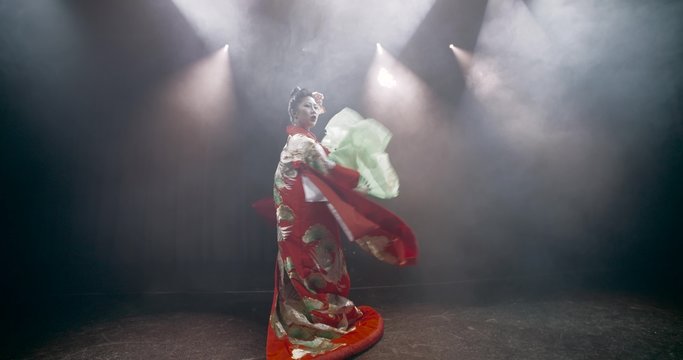 beautiful Japanese geisha posing for the camera with fans on stage, epic slow motion, smoke and dark background