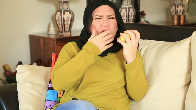 Muslim woman with a toothpain sitting on a sofa and eating pear