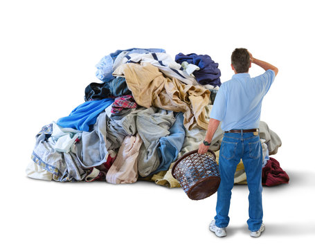 A distraught man with his hand to his head and a laundry basket in his hand is standing in front of a giant pile of dirty clothes overwhelmed by this household chore