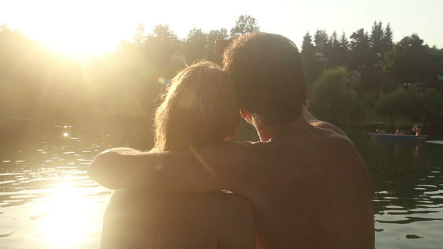 Couple in love taking selfies and kissing while sitting by the river at sunset
