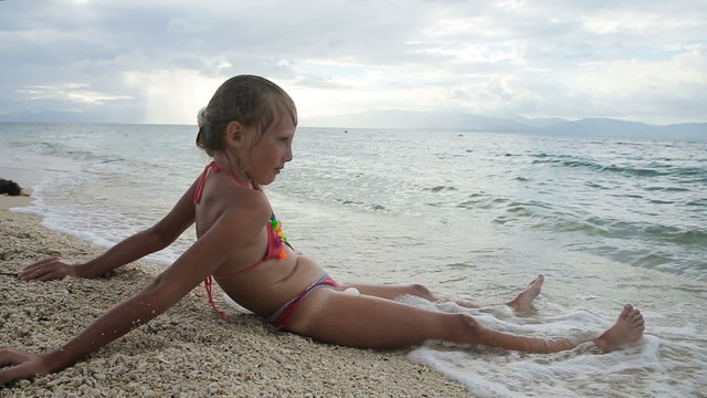 Young girl sitting on a tropical beach, watching the sea.A child resting on beach sand, the waves impinge on her legs.Sea wave, bare legs of a woman, a girl.Travel concept.Family,summer vacation