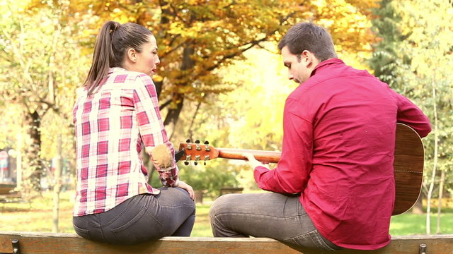 Couple sitting on park bench, man playing guitar while woman singing