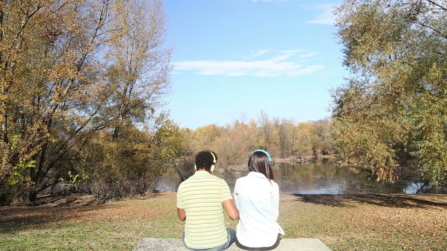 Man and woman listening to music using headphones while sitting by lake