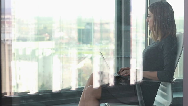 Businesswoman typing on laptop while sitting on chair behind glass wall