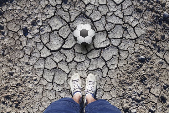 Man with white shoes standing on cracked dried soil ground with a soccer ball. Conceptual soccer ball game photo. Point of view man standing on cracked soil ground with a soccer ball.