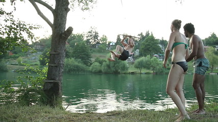 Handsome young man jumping off rope swing into river while happy friends cheer for him, in slow motion
