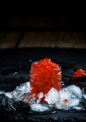 Delicious red caviar in a glass jar on crushed ice on black ston
