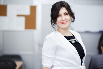 Successful attractive brunette with kind eyes standing in conference room and welcoming smile. Wearing white jacket, strict black dress jewelry. Portrait of cute young business woman posing.  