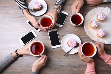 Four hands with smart phones holding  cups with tea, on wooden table background