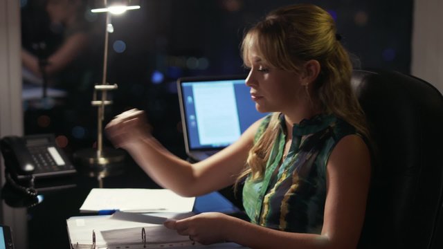 Beautiful blond business woman working overtime at night in executive office, reading documents and files on tablet pc. City lights are visible in background from a large window