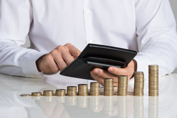 Businessman Using Calculator With Stacked Coins Arranged At Desk