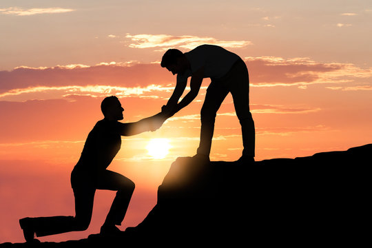 Man Assisting Male Friend In Climbing Rock During Sunset