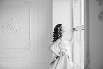 Bride looking out the window, she waits for the groom. Beautiful bride in white wedding dress standing in her bedroom and looking in window.
