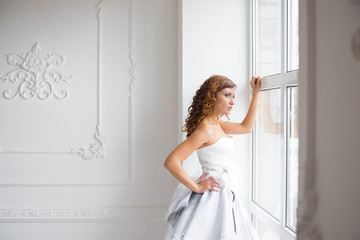 Bride looking out the window, she waits for the groom. Beautiful bride in white wedding dress standing in her bedroom and looking in window.
