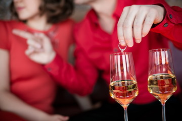 An engagement ring in a glass of champaign.  Young romantic couple in red.  Focus on a ring and glass of champaign. People are soft focus as a background. 