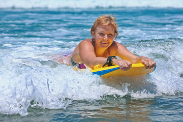 Joyful middle age woman - surfer with bodyboard surfing with fun on small sea waves. Active family lifestyle, people outdoor water sport lesson and swimming activity on summer vacation in ocean island