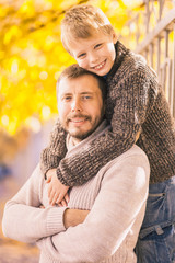 Obraz premium Portrait of cute little smiling kid hugging his daddy on sunny beautiful autumn day. Smiling people faces of boy and adult man looking at camera. Beautiful joyful family playing outside in fall park.