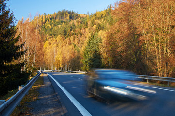 Asphalt road in a valley between forested mountains glowing autumn colors. Motion blur speeding car. Sunny autumn day with blue sky.