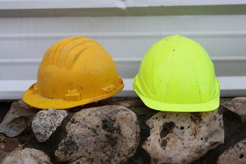 Two Work helmets of a construction worker on a rock fence in the construction site. Yellow and orange wet safety helmets of a construction worker in front of a corrugated iron fence in a rainy day.