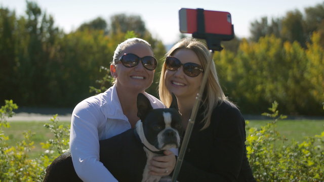 Two Pretty Young Women with Dog Taking Photos with Smart Phone on Selfie Stick. Blond and Brunette Taking Cell Phone Selfies with Boston Terrier on Sunny Day