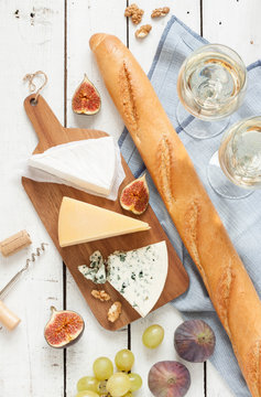 Different kinds of cheeses (brie, parmesan, blue cheese) baguette, two glasses of white wine, figs and grapes. White wooden table as background. Romantic french picnic scenery captured from above.
