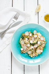 Salad - grilled zucchini 'tagliatelle', feta cheese, radish and red onion slices, roasted sunflower seeds and vinaigrette dressing on blue plate from above. White rustic planked table as background.