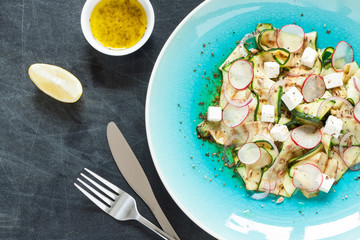 Salad - grilled zucchini 'tagliatelle', feta cheese, radish and red onion slices, roasted sunflower seeds and vinaigrette dressing on blue plate from above. Black chalkboard as background.