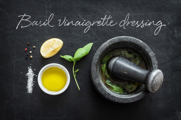 Basil vinaigrette dressing - recipe ingredients on black chalkboard background from above. Fresh basil leaves, lemon, olive oil, salt, pepper and mortar. Kitchen poster layout.