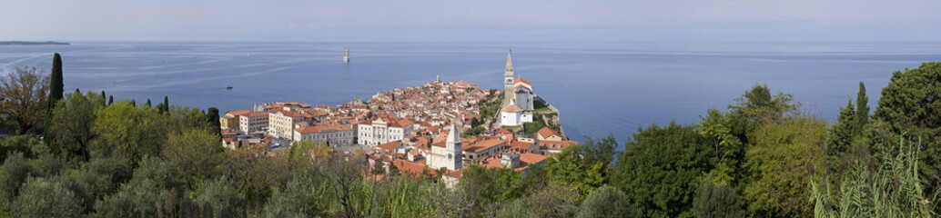 Fototapeta na wymiar Piran (Italian: Pirano) old town in Slovenia, Gulf of Piran on the Adriatic Sea..Panoramic view of the city. Natural green environment, blue sky, red roofs of houses and Church of Saint George .