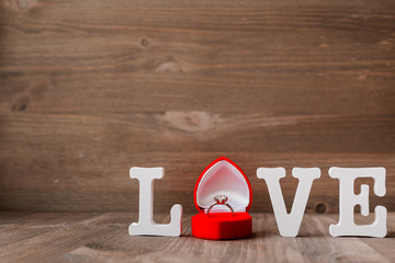 Word LOVE and diamond engagement ring in red gift box on wooden background.