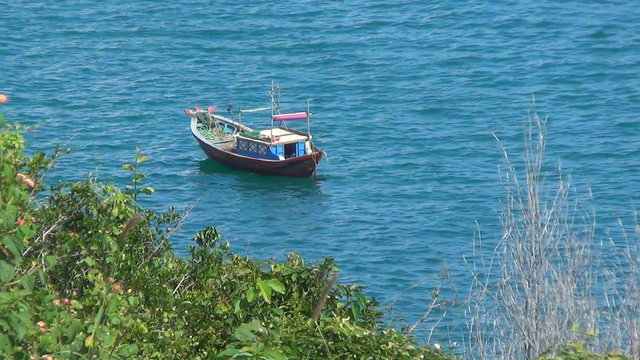 small wooden boat rocking on the waves. Bay is surrounded by greenery. Blue Sea has a slight wave.