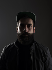 Serious unconventional bearded man wearing baseball cap staring at camera. High contrast low key dark shadow portrait over black background.