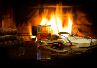 Glass of alcoholic drink and antique books in front of warm fireplace. Magical relaxed cozy...