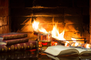 Glass of alcoholic drink and antique books in front of warm fireplace. Magical relaxed cozy...