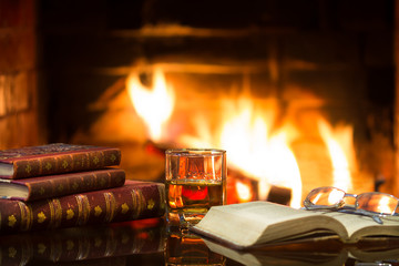 Glass of alcoholic drink and antique books in front of warm fireplace. Magical relaxed cozy atmosphere near fire