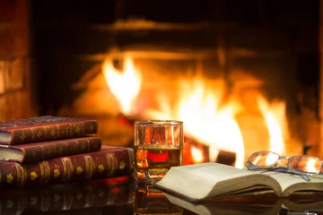Poster de jardin Bar Glass of alcoholic drink and antique books in front of warm fireplace. Magical relaxed cozy atmosphere near fire