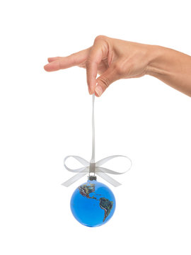 Photograph of a female hand dangling a glass Christmas ornament of the earth suspended on a white ribbon isolated on a white background.