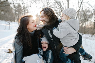 Happy young hipster family having fun, bowl, rising up, piggyback ride their children and relax in park on winter sunset
