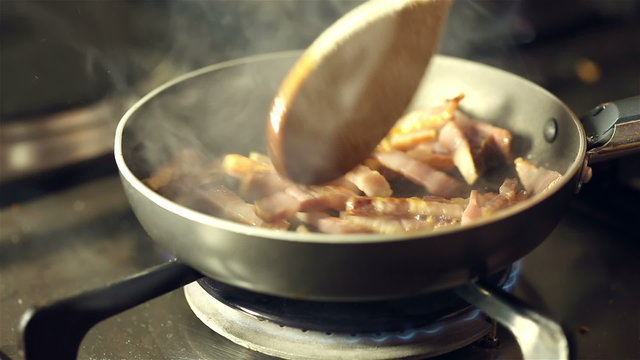 Small pieces of pancetta frying in the skilet and stirring with wooden spoon on gas stove in preparation of spaghetti carbonara