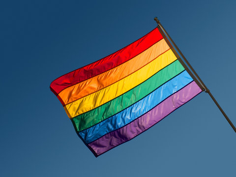 Photograph of a rainbow flag against a clear blue sky.