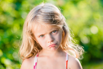 Close-up portrait of displeased blond little girl with pursed lips. Sunny summer day in beautiful park