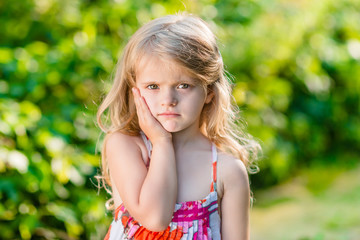 Sad little girl with long blond hair suffering from toothache. Sunny summer day in beautiful green park