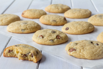 Homemade Chocolate Chip Cookies on Baking Paper with One Partly Eaten on White Wooden Table