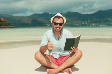 man sitting on the beach with book showing success sign