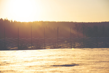 Handcrafted wooden fence against a beautiful sunset. Some snow is on the ground on a sunny winter day in Finland. Image has a vintage and flare effect applied.