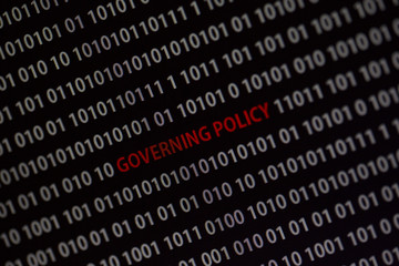 'Governing policy' text in the middle of the computer screen surrounded by numbers zero and one. Image is taken in a small angle.
